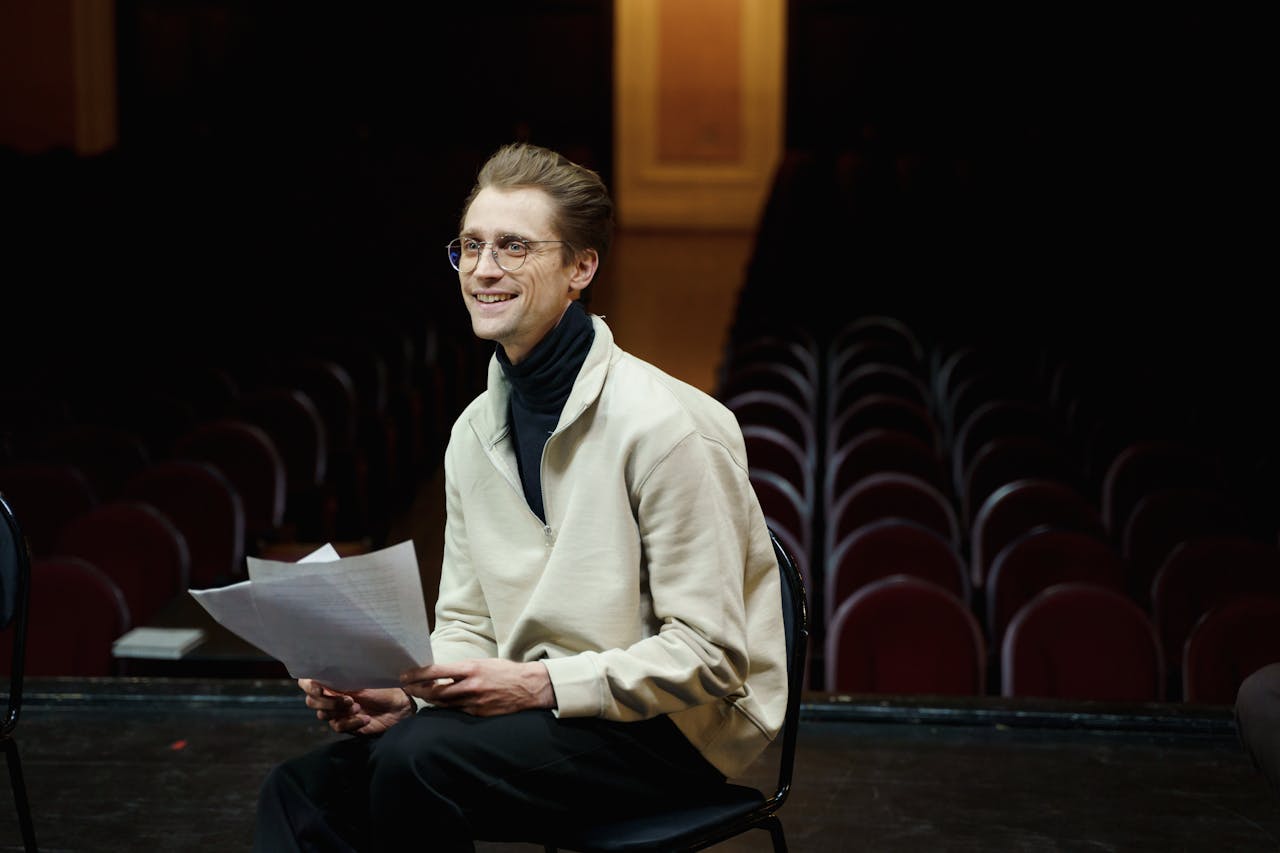 Smiling actor rehearsing script alone in empty theater auditorium, seated on stage.