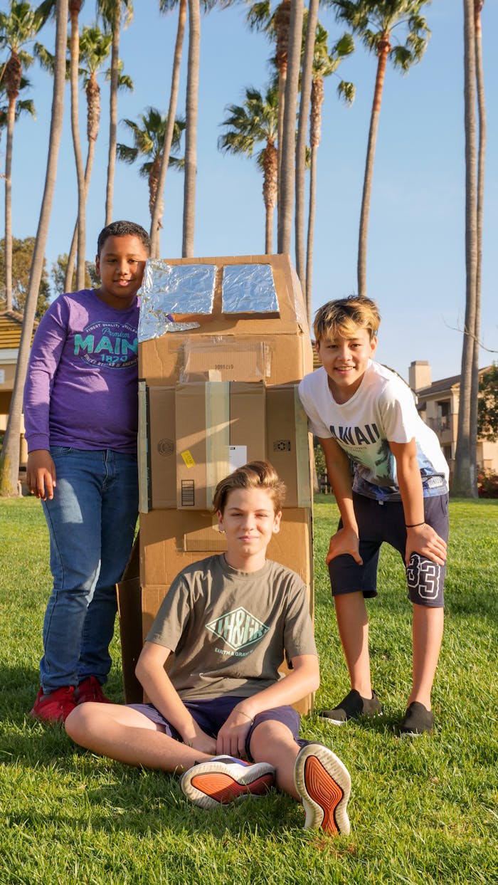 Three kids enjoy outdoor playtime with a handmade cardboard rocket on a sunny day.