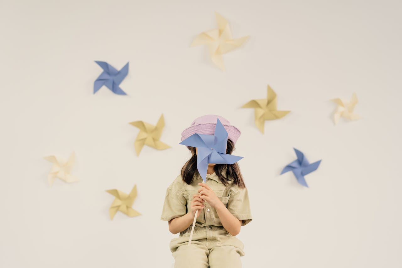 A young girl holding a blue pinwheel, surrounded by colorful paper pinwheels on a wall.