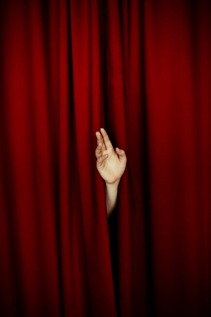 A hand appears between rich red velvet curtains, evoking a theatrical performance.