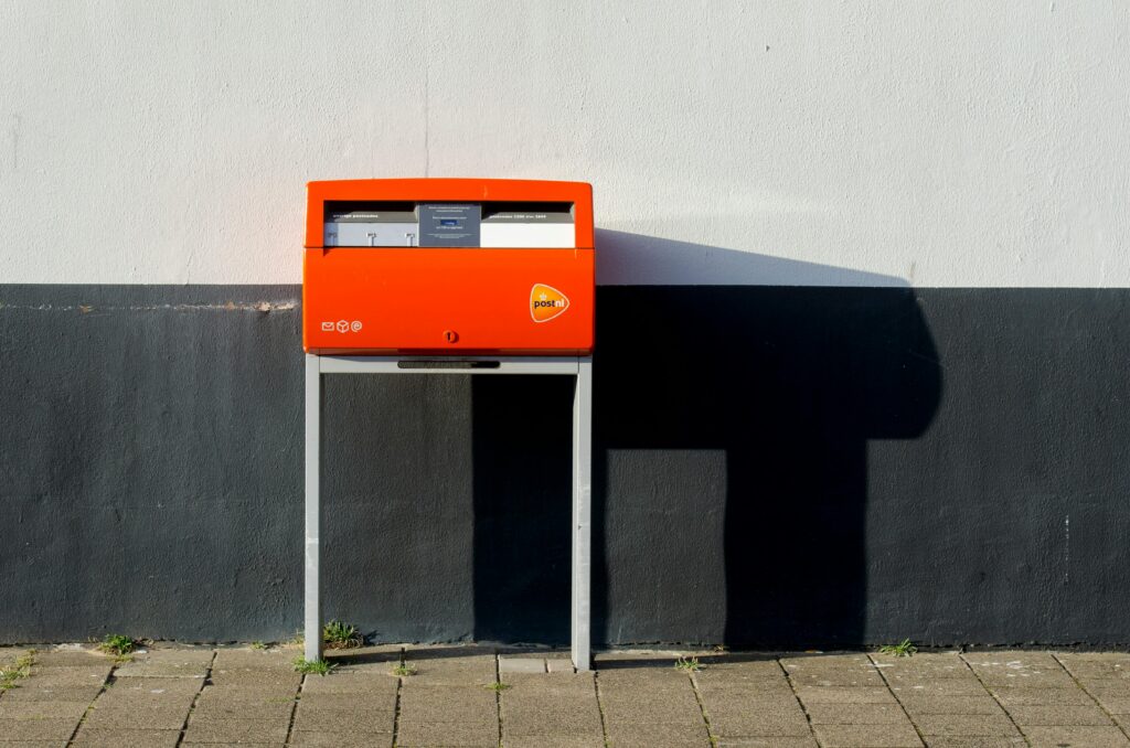 A vibrant PostNL mailbox set against a city wall casting a shadow in daylight.